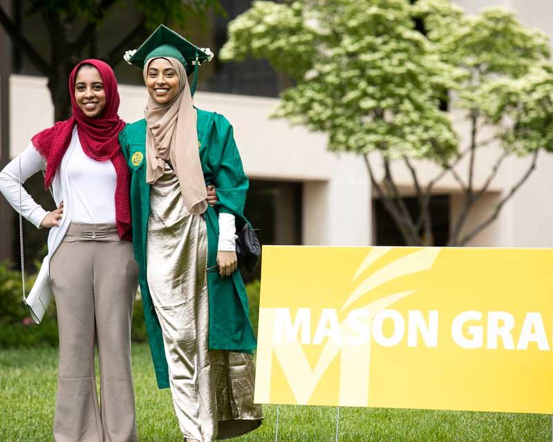 Friends pose together at the Patriot Procession wearing graduation regalia and standing near environmental graphics celebrating the event.